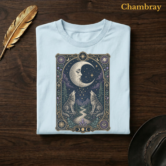 Moon tarot tee in Chambray, a soft pale blue garment-dyed shirt folded flat on dark walnut wood beside a gold feather and a leather hat, the weeping moon and crystal path design on the chest, labelled Chambray.