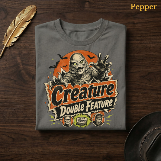 Creature Double Feature tee in Pepper, a warm grey-charcoal garment-dyed shirt folded flat on dark walnut wood beside a gold feather and a leather hat, the pulp horror swamp monster design centred on the chest, labelled Pepper.