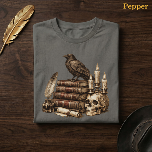 Macabre poetry tee in Pepper, a warm grey-charcoal garment-dyed shirt folded flat on dark walnut wood beside a gold feather and a leather hat, the Poe's Study raven design centred on the chest, labelled Pepper.