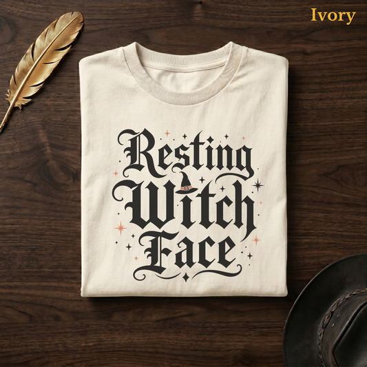 Resting Witch Face tee in Ivory, folded flat on dark walnut wood beside a gold feather and a leather hat, the blackletter witch hat lettering design on the chest, labelled Ivory.