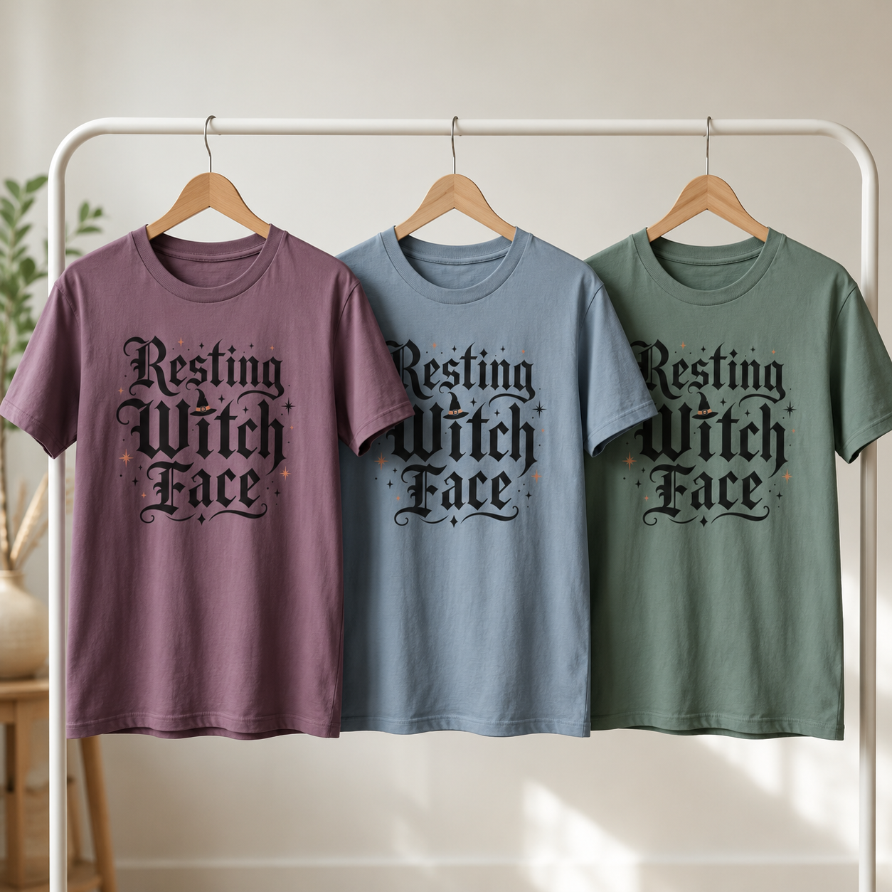 Resting Witch Face tee shown in three colorways on wooden hangers on a clothing rack: Berry, Blue Jean, and Blue Spruce, each with the blackletter witch design centred on the chest.