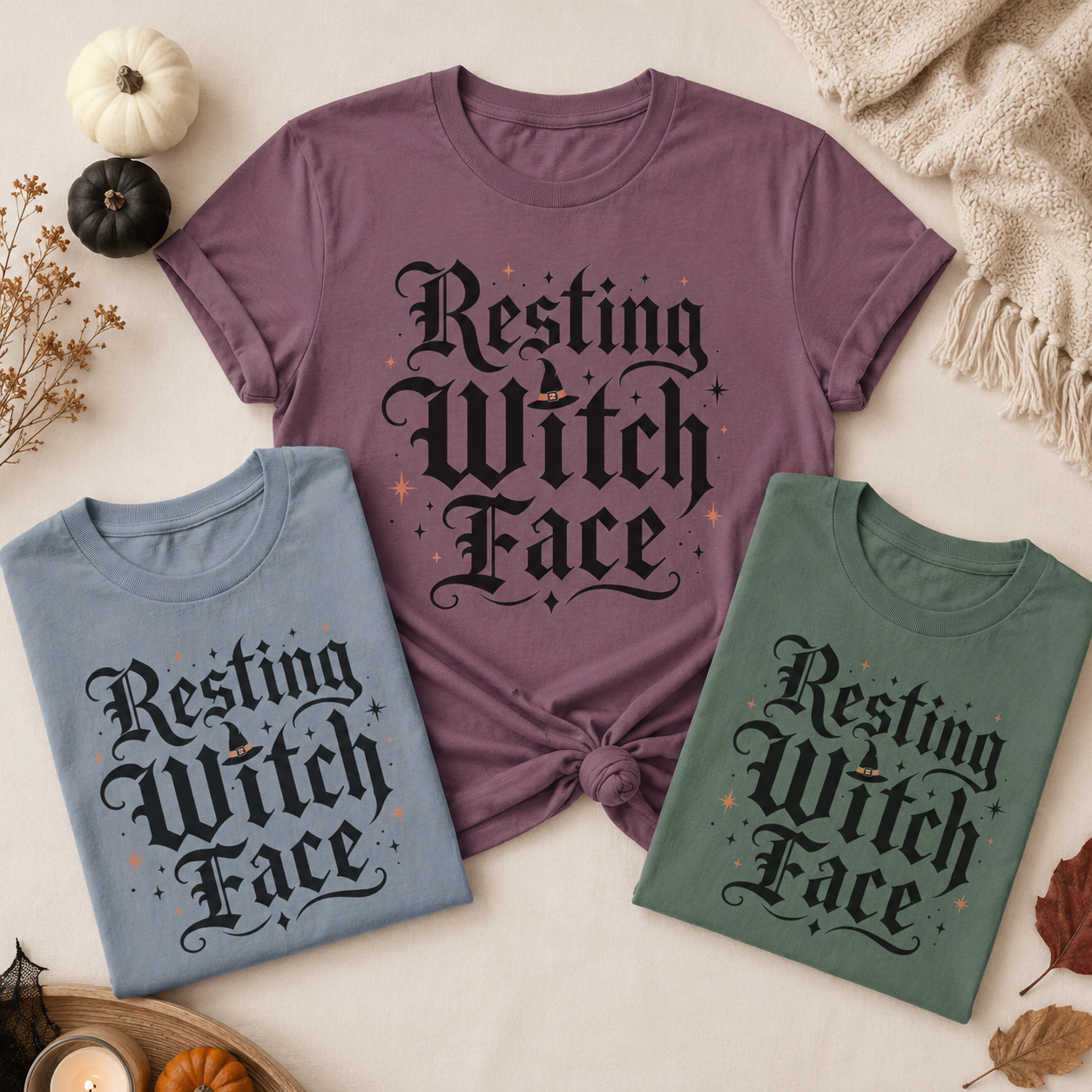 Sassy halloween tee shown in three settings: a knotted Berry tee, a folded Blue Jean tee, and a folded Blue Spruce tee, styled with mini pumpkins, dried flowers, a lit candle, and a knit throw, each printed with the Resting Witch Face design.