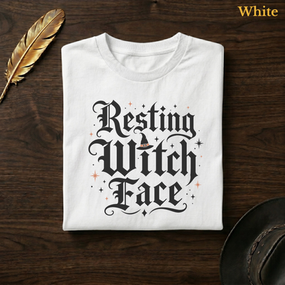Resting Witch Face tee in White, folded flat on dark walnut wood beside a gold feather and a leather hat, the full blackletter witch hat lettering design printed on the chest, labelled White.
