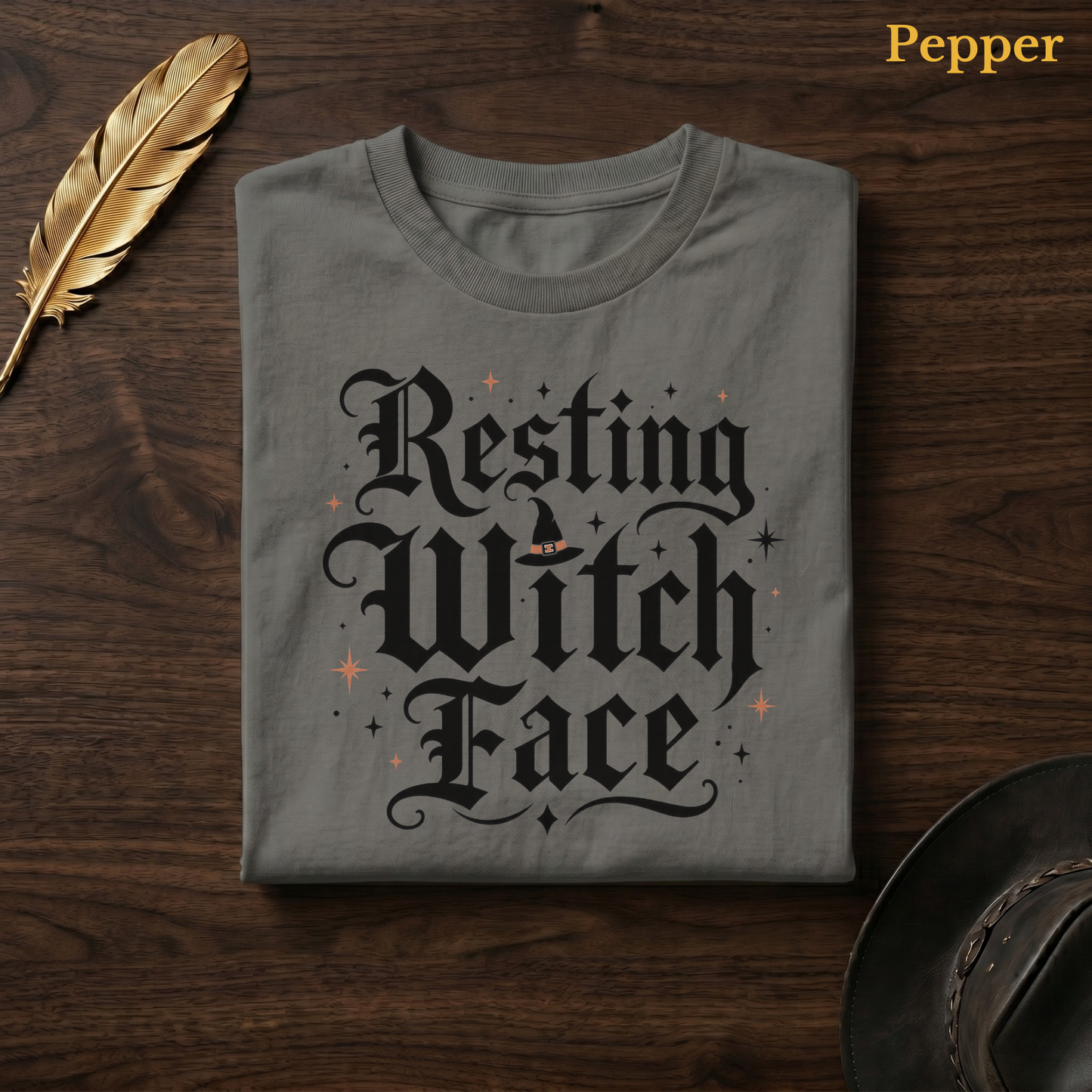Resting Witch Face tee in Pepper, a warm grey-charcoal garment-dyed shirt folded flat on dark walnut wood beside a gold feather and a leather hat, the funny witch lettering design centred on the chest, labelled Pepper.