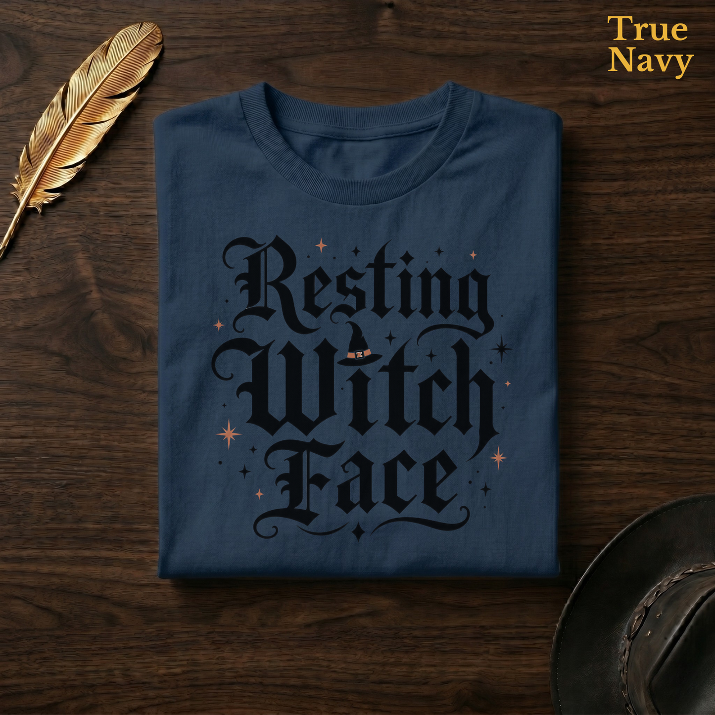 Resting Witch Face tee in True Navy, a deep navy garment-dyed shirt folded flat on dark walnut wood beside a gold feather and a leather hat, the blackletter witch design on the chest, labelled True Navy.