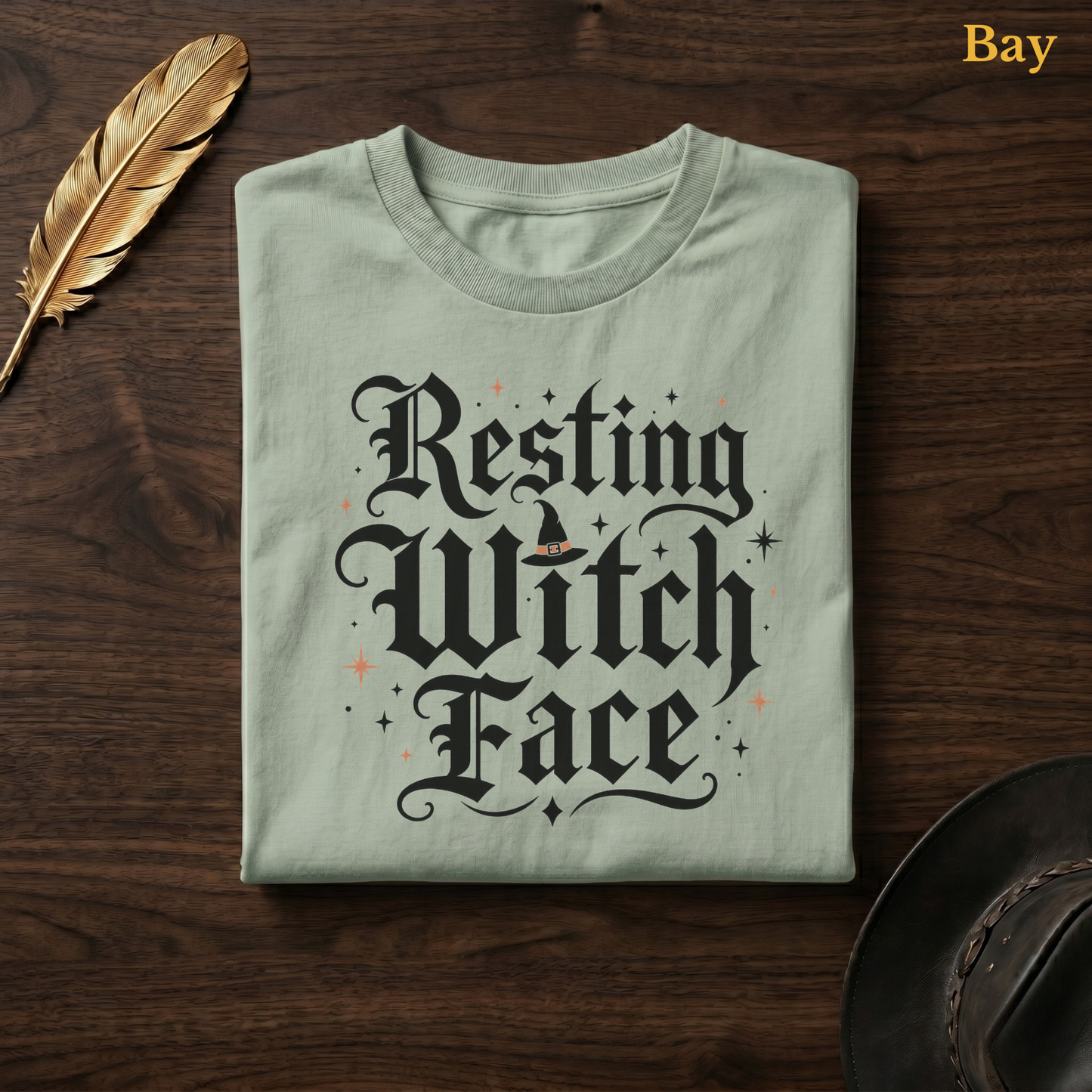 Resting Witch Face tee in Bay, a soft sage-green garment-dyed shirt folded flat on dark walnut wood beside a gold feather and a leather hat, the blackletter witch design printed on the chest, labelled Bay.