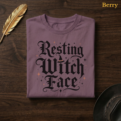 Resting Witch Face tee in Berry, a muted mauve-plum garment-dyed shirt folded flat on dark walnut wood beside a gold feather and a leather hat, the funny witch lettering design on the chest, labelled Berry.