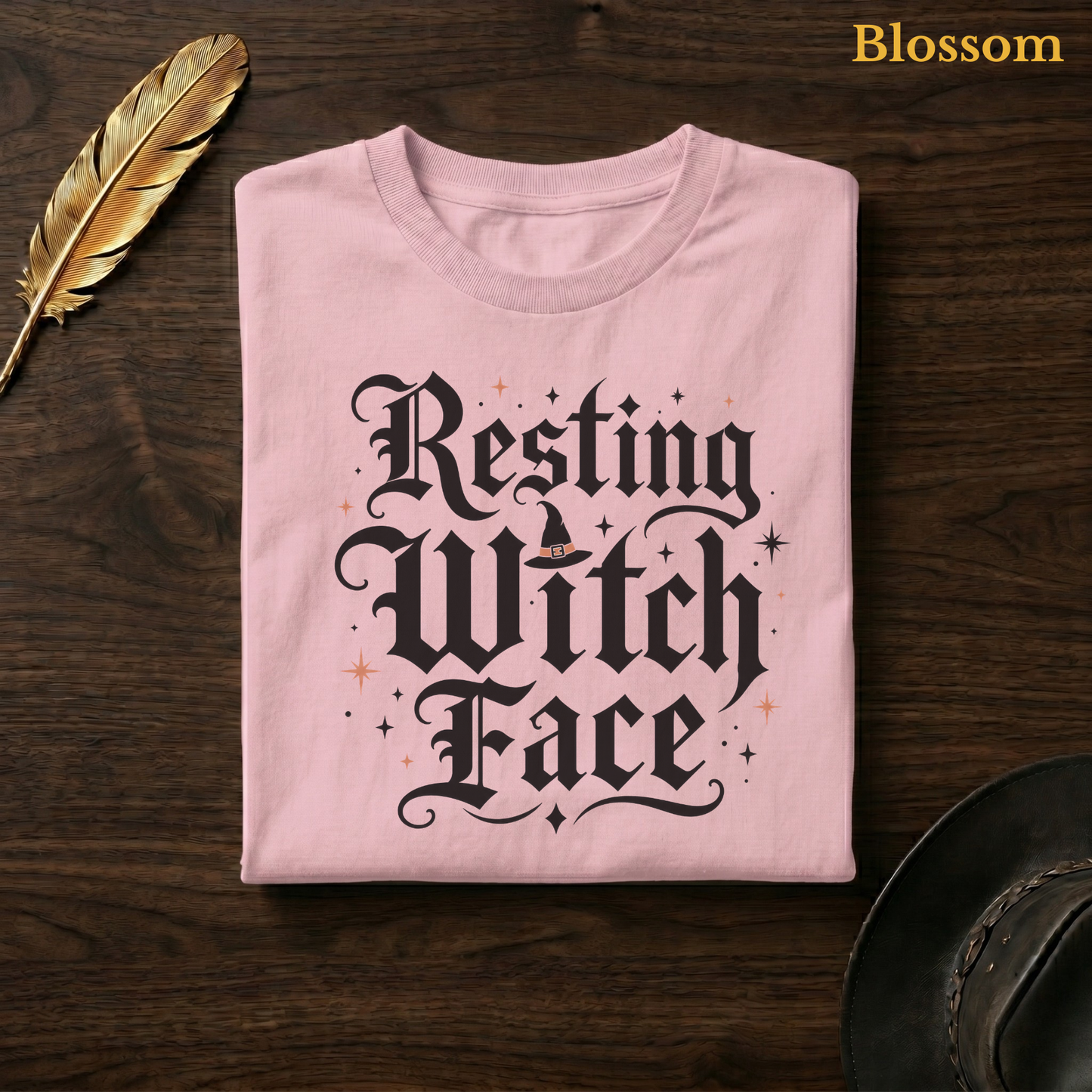 Resting Witch Face tee in Blossom, a soft pastel pink garment-dyed shirt folded flat on dark walnut wood beside a gold feather and a leather hat, the witch hat lettering design on the chest, labelled Blossom.