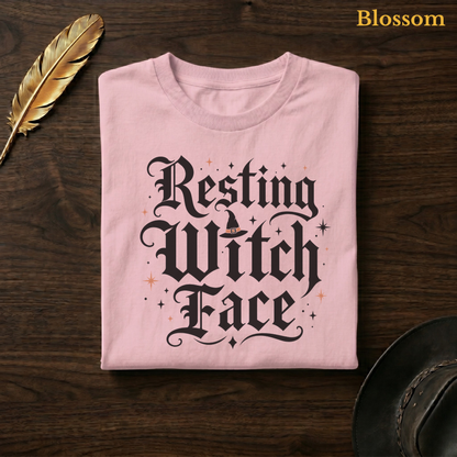 Resting Witch Face tee in Blossom, a soft pastel pink garment-dyed shirt folded flat on dark walnut wood beside a gold feather and a leather hat, the witch hat lettering design on the chest, labelled Blossom.