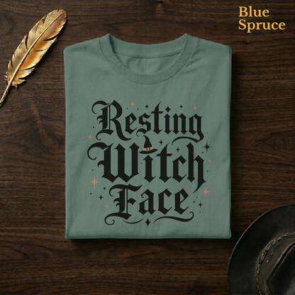 Resting Witch Face tee in Blue Spruce, a deep teal-green garment-dyed shirt folded flat on dark walnut wood beside a gold feather and a leather hat, the blackletter witch design on the chest, labelled Blue Spruce.
