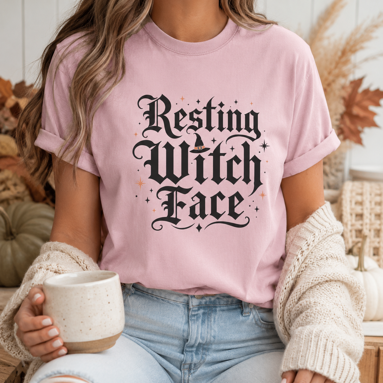 Witchy humor tee in Blossom worn by a woman holding a ceramic coffee mug, styled with pumpkins, dried leaves, and a cozy knit cardigan, the Resting Witch Face design centred on the chest.