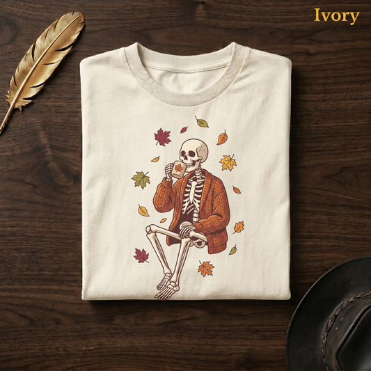 Sweater Weather Skeleton tee in Ivory, folded flat on dark walnut wood beside a gold feather and a leather hat, the skeleton, mug, and falling leaves design on the chest, labelled Ivory.
