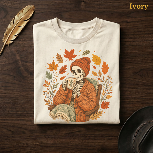 Sweater Weather Skeleton tee in Ivory, folded flat on dark walnut wood beside a gold feather and a leather hat, the beanie skeleton, mug, and wreath design on the chest, labelled Ivory.