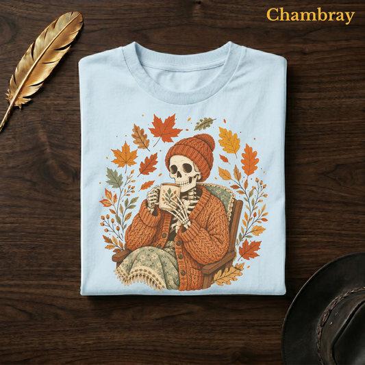Sweater Weather Skeleton tee in Chambray, a soft pale blue garment-dyed shirt folded flat on dark walnut wood beside a gold feather and a leather hat, the cardigan skeleton and wreath design on the chest, labelled Chambray.
