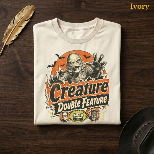 Creature Double Feature tee in Ivory, folded flat on dark walnut wood beside a gold feather and a leather hat, the vintage monster and title-card design on the chest, labelled Ivory.
