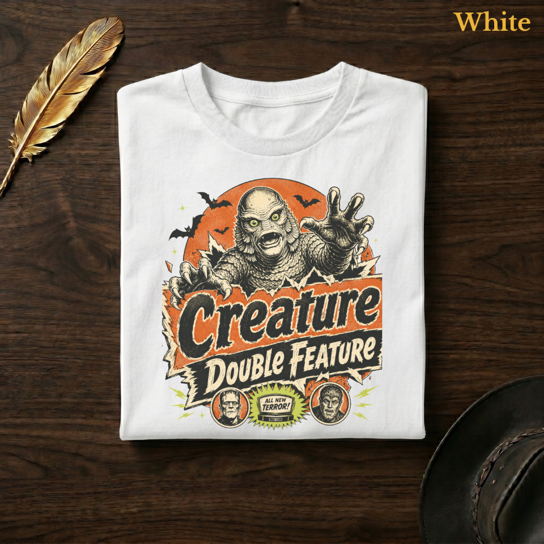 Creature Double Feature tee in White, folded flat on dark walnut wood beside a gold feather and a leather hat, the full-colour swamp monster, title banner, and monster medallions design printed on the chest, labelled White.
