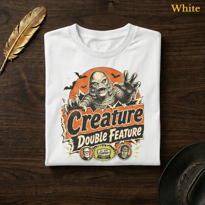 Creature Double Feature tee in White, folded flat on dark walnut wood beside a gold feather and a leather hat, the full-colour swamp monster, title banner, and monster medallions design printed on the chest, labelled White.

