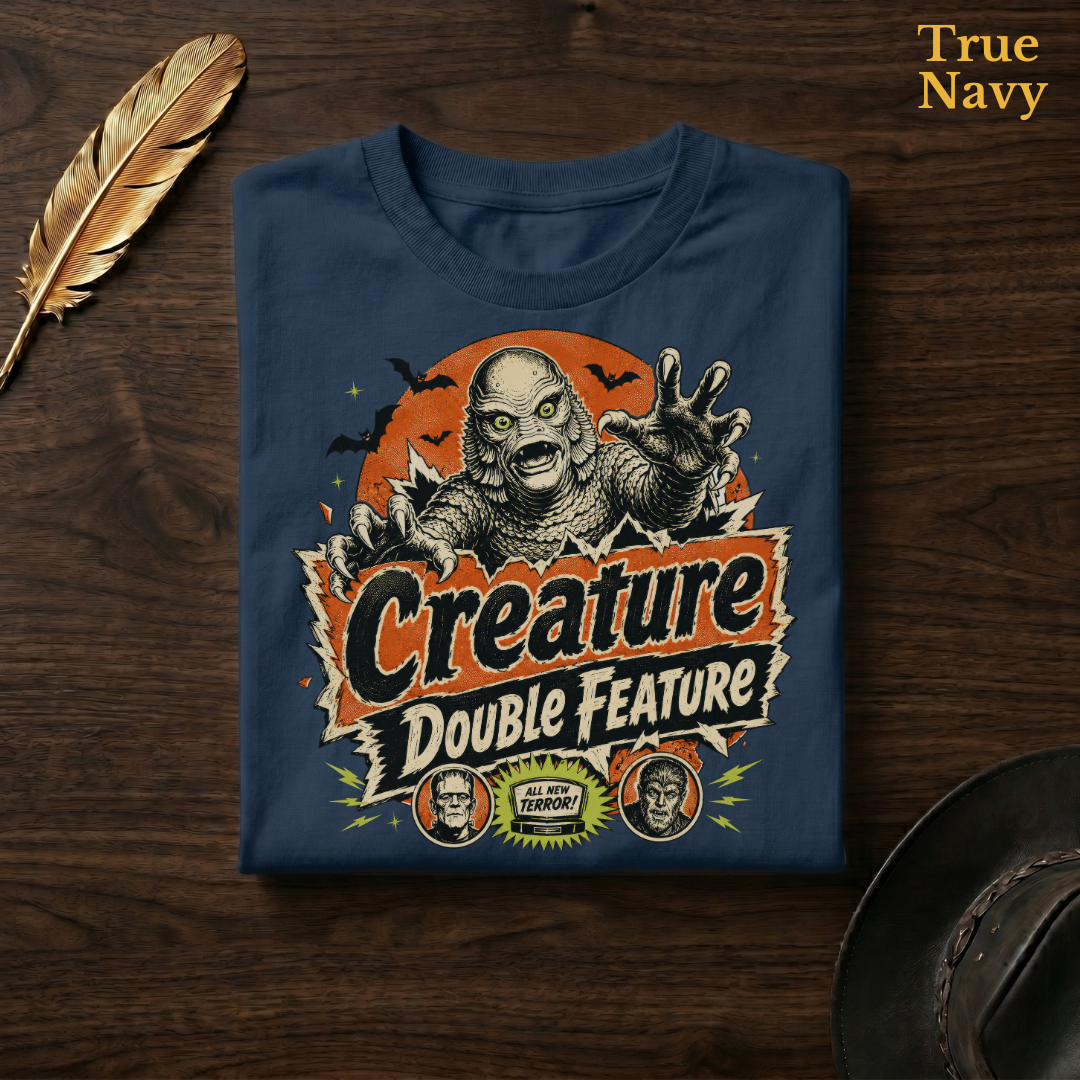 Creature Double Feature tee in True Navy, a deep navy garment-dyed shirt folded flat on dark walnut wood beside a gold feather and a leather hat, the retro monster title-card design on the chest, labelled True Navy.
