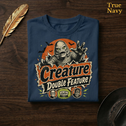 Creature Double Feature tee in True Navy, a deep navy garment-dyed shirt folded flat on dark walnut wood beside a gold feather and a leather hat, the retro monster title-card design on the chest, labelled True Navy.
