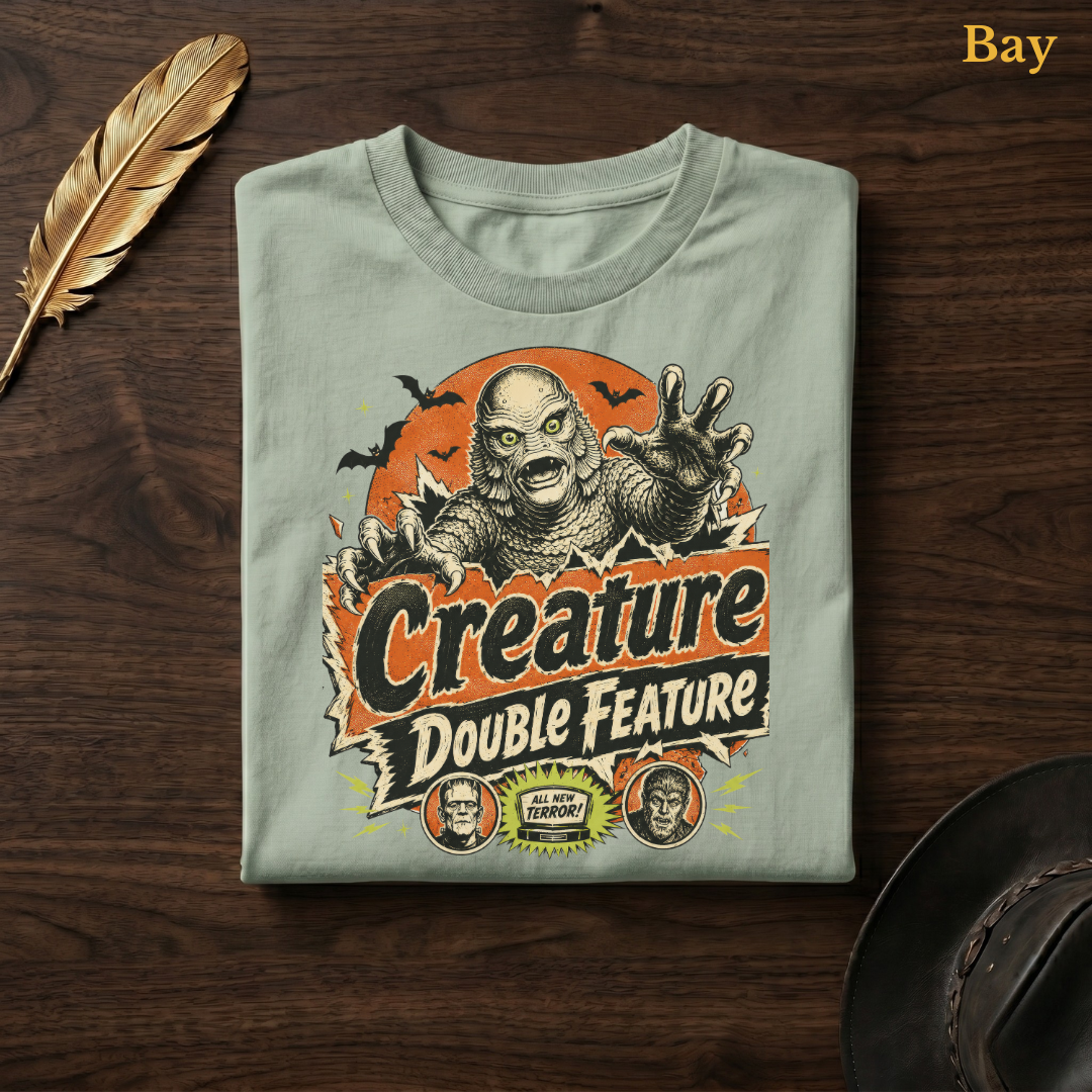 Creature Double Feature tee in Bay, a soft sage-green garment-dyed shirt folded flat on dark walnut wood beside a gold feather and a leather hat, the retro swamp monster design printed on the chest, labelled Bay.
