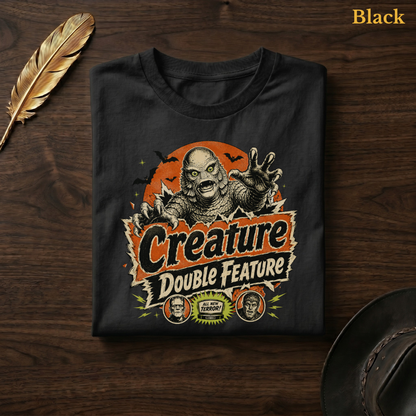 Creature Double Feature tee in Black, folded flat on dark walnut wood beside a gold feather and a leather hat, the pulp horror swamp monster and title-card design printed on the chest, labelled Black.

