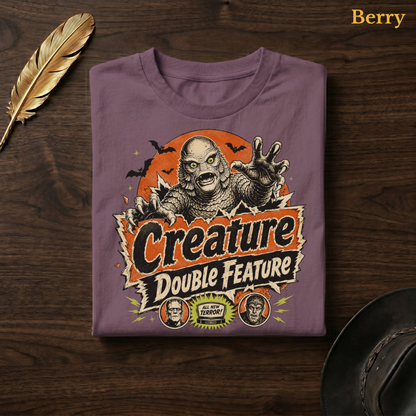 Creature Double Feature tee in Berry, a muted mauve-plum garment-dyed shirt folded flat on dark walnut wood beside a gold feather and a leather hat, the vintage horror title-card design on the chest, labelled Berry.
