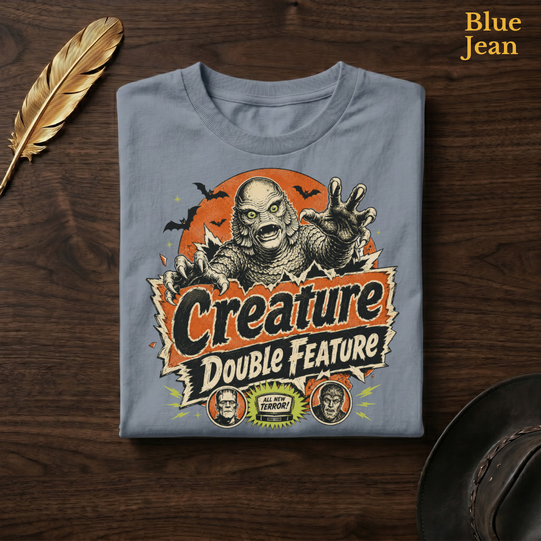 Creature Double Feature tee in Blue Jean, a faded denim-blue garment-dyed shirt folded flat on dark walnut wood beside a gold feather and a leather hat, the b movie horror design on the chest, labelled Blue Jean.
