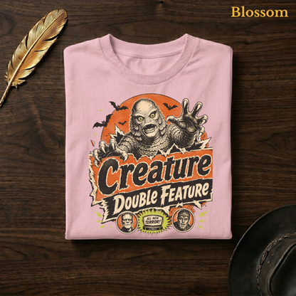 Creature Double Feature tee in Blossom, a soft pastel pink garment-dyed shirt folded flat on dark walnut wood beside a gold feather and a leather hat, the retro monster design on the chest, labelled Blossom.
