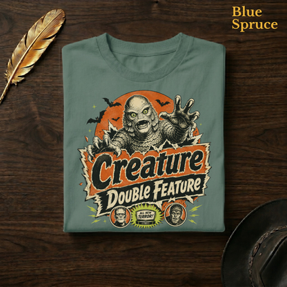 Creature Double Feature tee in Blue Spruce, a deep teal-green garment-dyed shirt folded flat on dark walnut wood beside a gold feather and a leather hat, the swamp monster title-card design on the chest, labelled Blue Spruce.

