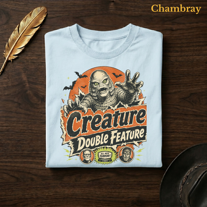 Creature Double Feature tee in Chambray, a soft pale blue garment-dyed shirt folded flat on dark walnut wood beside a gold feather and a leather hat, the retro horror poster design on the chest, labelled Chambray.
