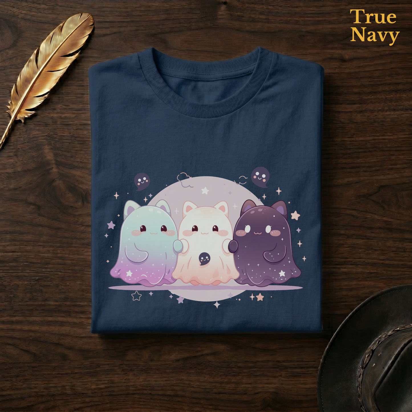 Celestial cat tee in True Navy, a deep navy garment-dyed shirt folded flat on dark walnut wood beside a gold feather and a leather hat, the moonlit cat-ghost trio design on the chest, labelled True Navy.
