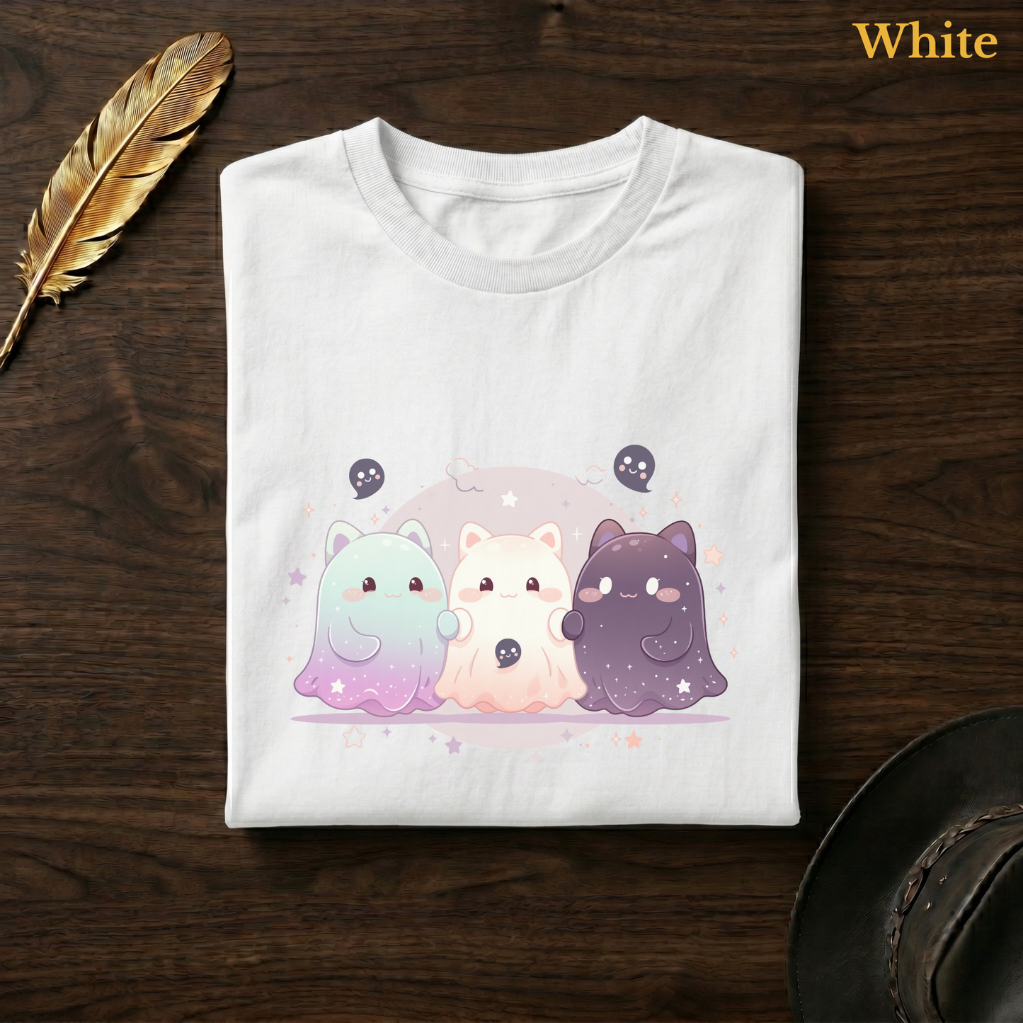 Cosmic kawaii shirt in White, folded flat on dark walnut wood beside a gold feather and a leather hat, the full-colour galaxy cat-ghost trio and moon design printed on the chest, labelled White.
