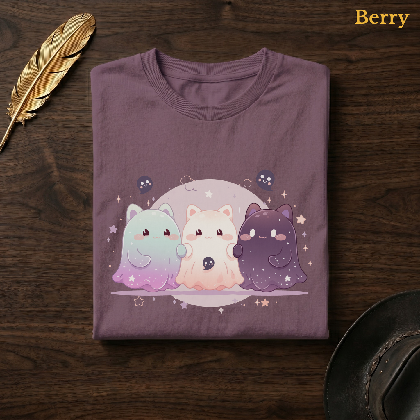 Ghoul Friends tee in Berry, a muted mauve-plum garment-dyed shirt folded flat on dark walnut wood beside a gold feather and a leather hat, the starry cat-ghost design on the chest, labelled Berry.
