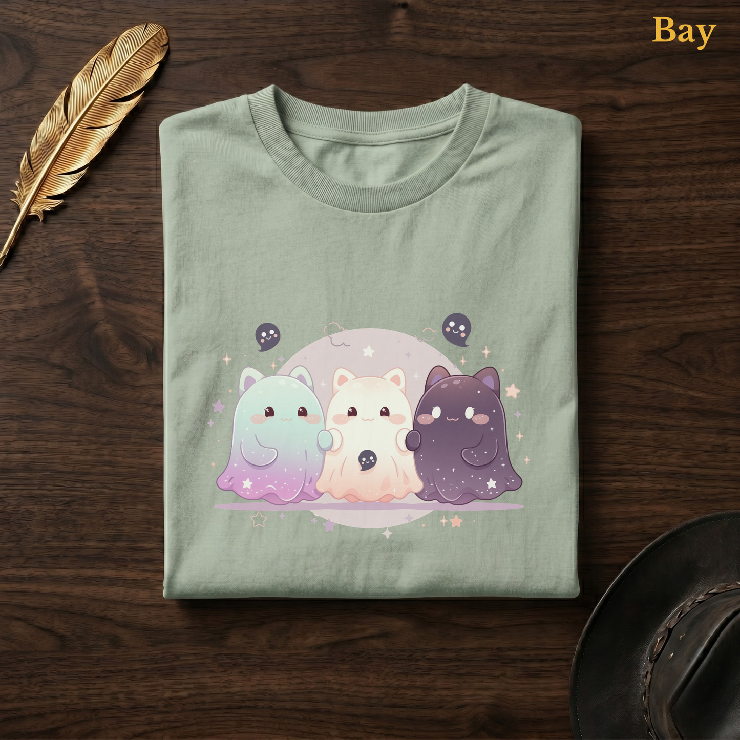 Ghoul Friends tee in Bay, a soft sage-green garment-dyed shirt folded flat on dark walnut wood beside a gold feather and a leather hat, the galaxy cat-ghost trio design printed on the chest, labelled Bay.
