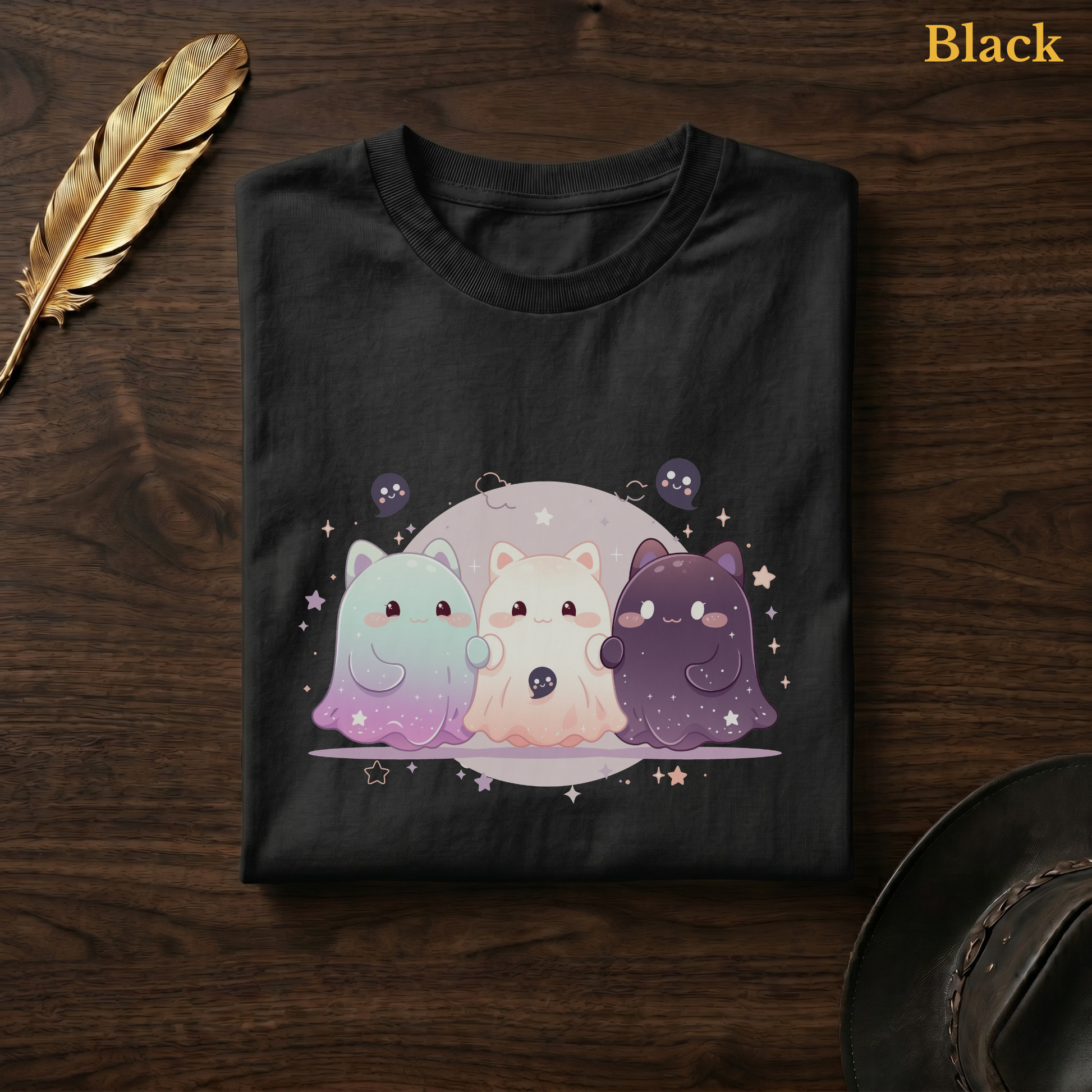 Ghoul Friends tee in Black, folded flat on dark walnut wood beside a gold feather and a leather hat, the celestial gradient cat-ghost trio design printed on the chest, labelled Black.

