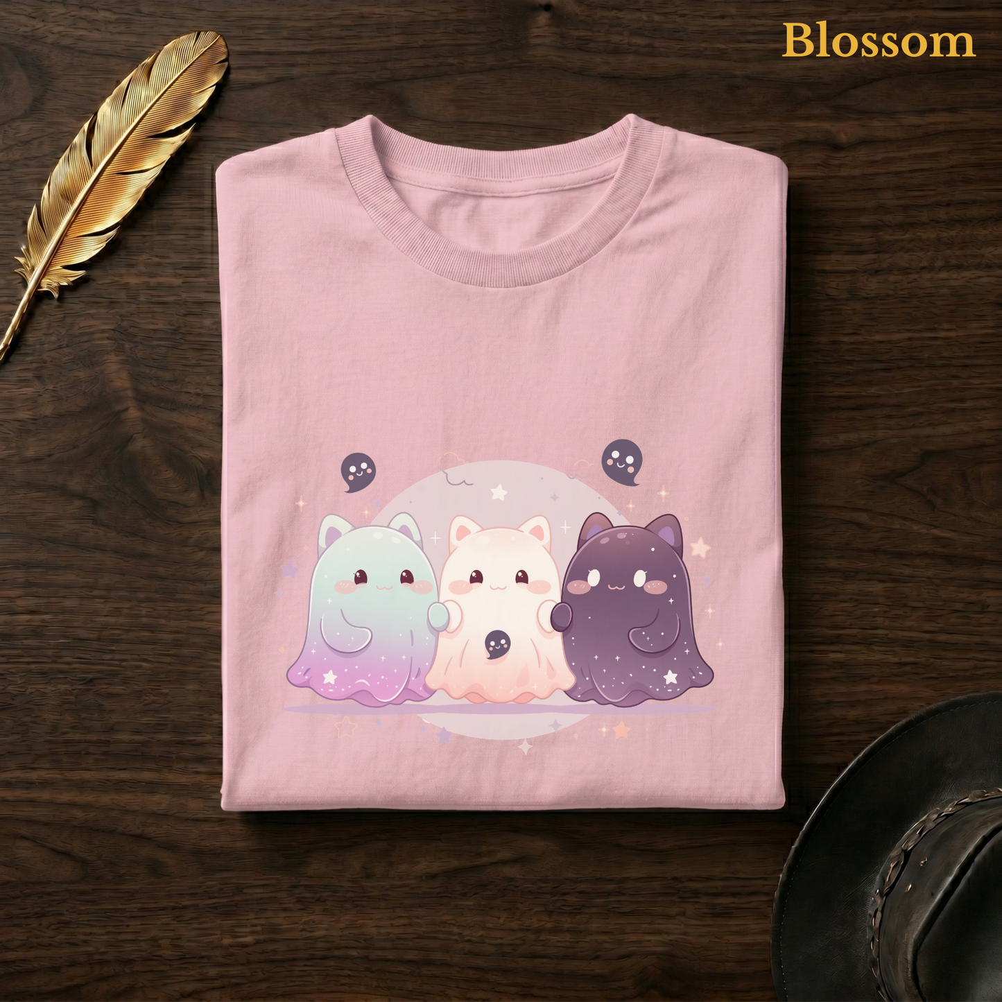 Ghoul Friends tee in Blossom, a soft pastel pink garment-dyed shirt folded flat on dark walnut wood beside a gold feather and a leather hat, the galaxy ghost design on the chest, labelled Blossom.
