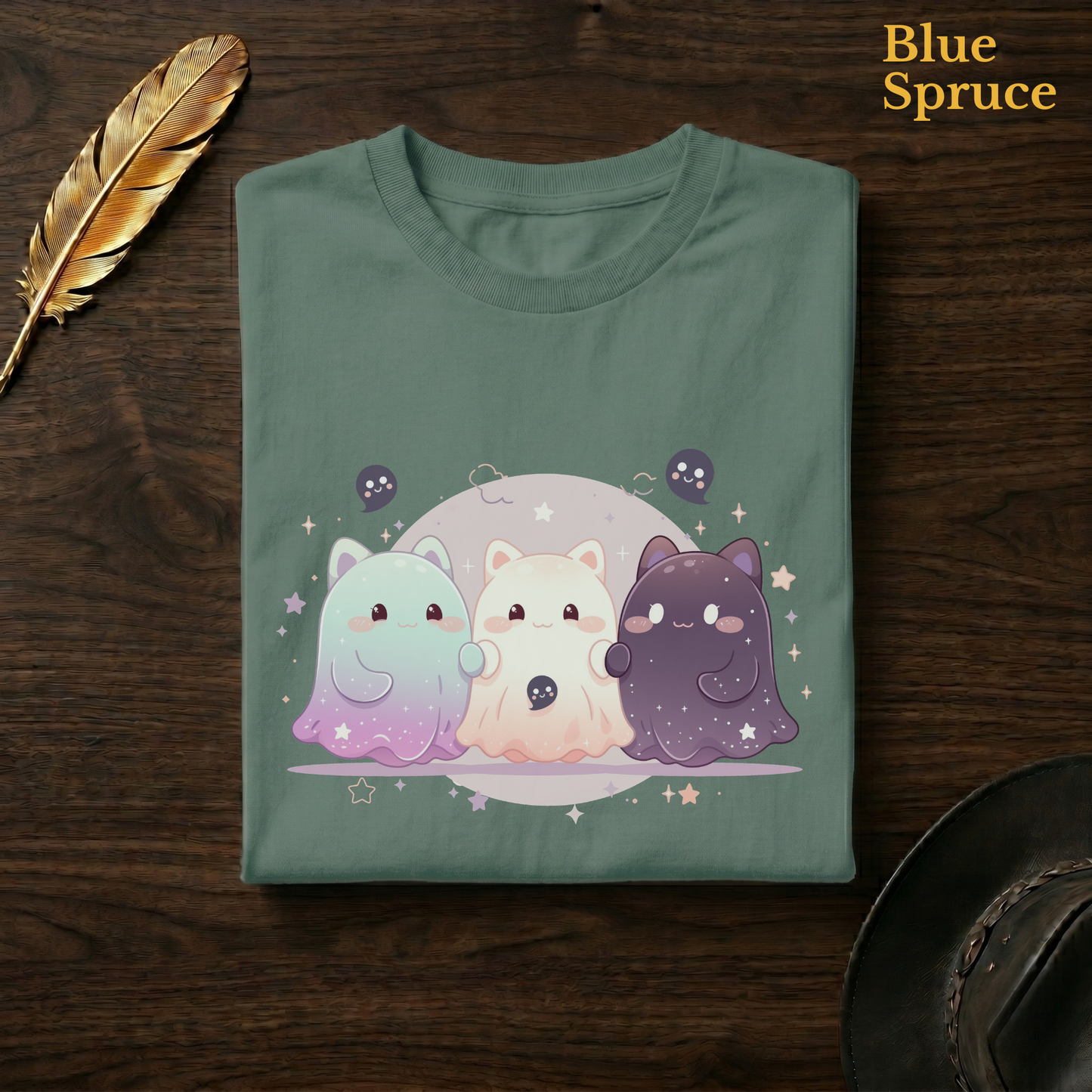 Night sky ghost tee in Blue Spruce, a deep teal-green garment-dyed shirt folded flat on dark walnut wood beside a gold feather and a leather hat, the celestial cat-ghost design on the chest, labelled Blue Spruce.
