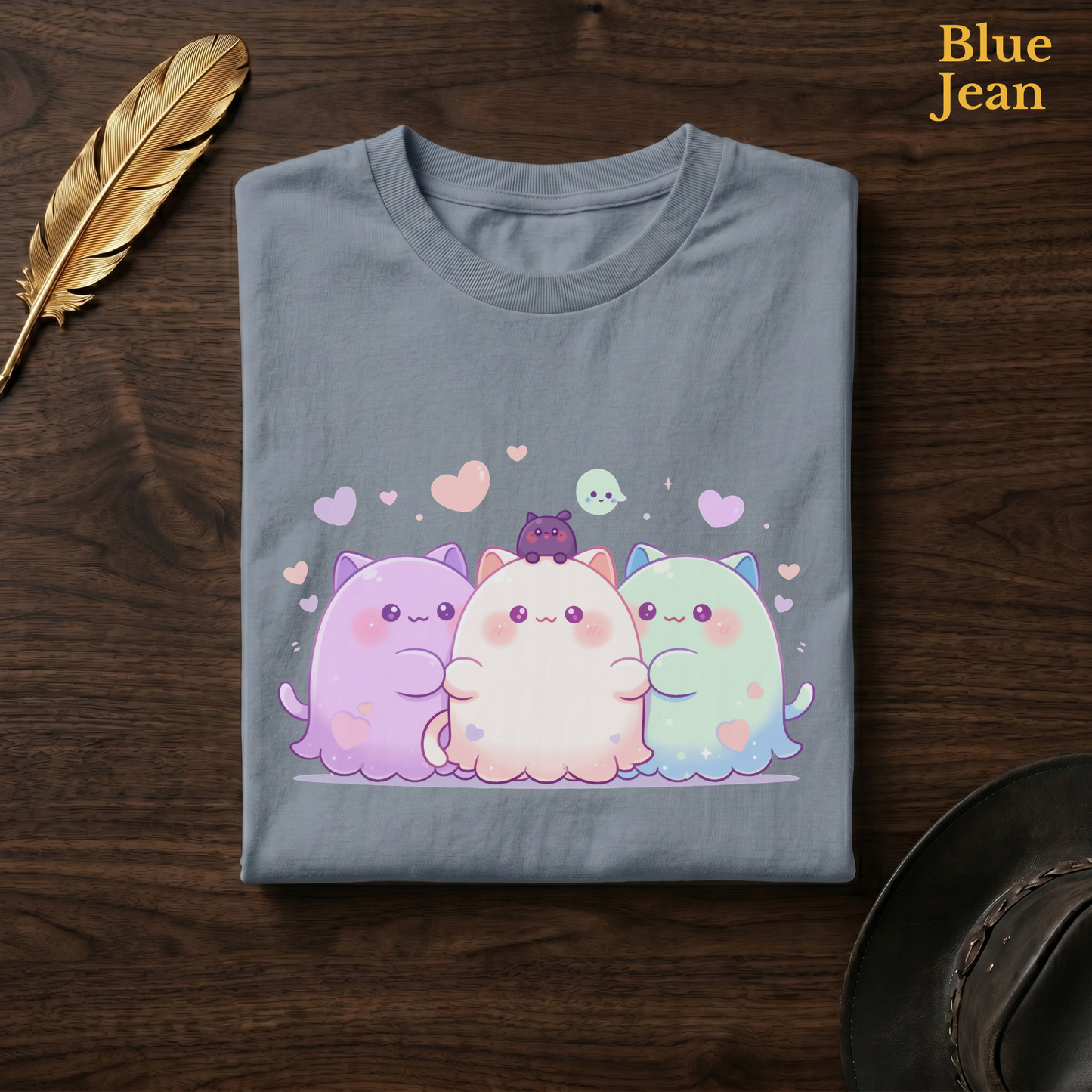 Cat ghost trio gift tee in Blue Jean, a faded denim-blue garment-dyed shirt folded flat on dark walnut wood beside a gold feather and a leather hat, the Ghoul Friends blob design on the chest, labelled Blue Jean.