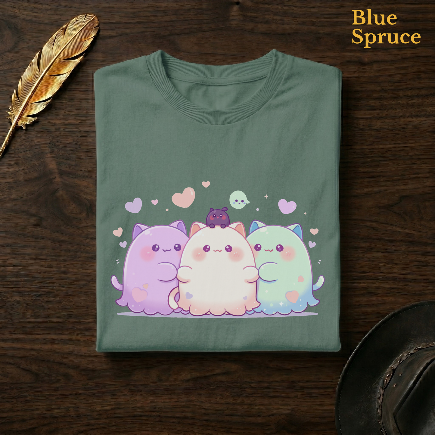 Plushie ghost tee in Blue Spruce, a deep teal-green garment-dyed shirt folded flat on dark walnut wood beside a gold feather and a leather hat, the kawaii cat-ghost hug design on the chest, labelled Blue Spruce.
