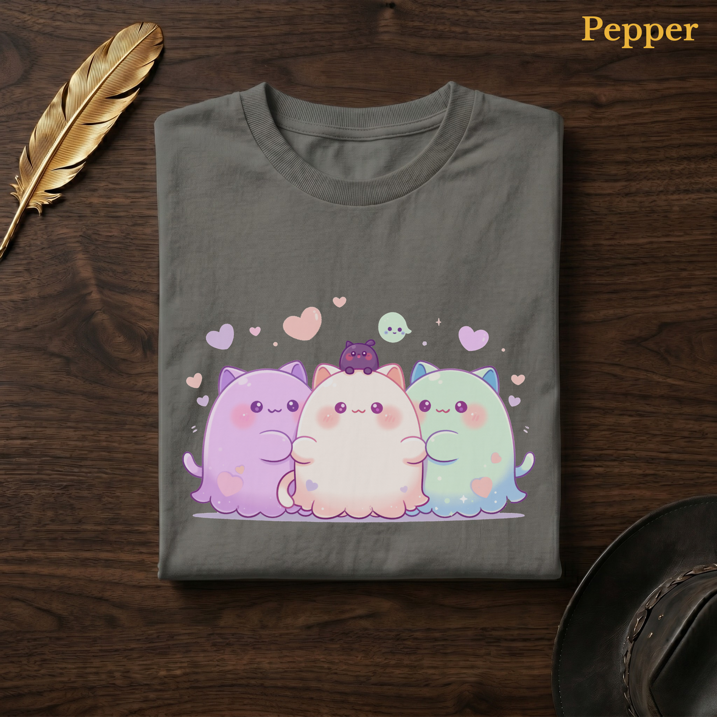Halloween bff tee in Pepper, a warm grey-charcoal garment-dyed shirt folded flat on dark walnut wood beside a gold feather and a leather hat, the Ghoul Friends blob design centred on the chest, labelled Pepper.
