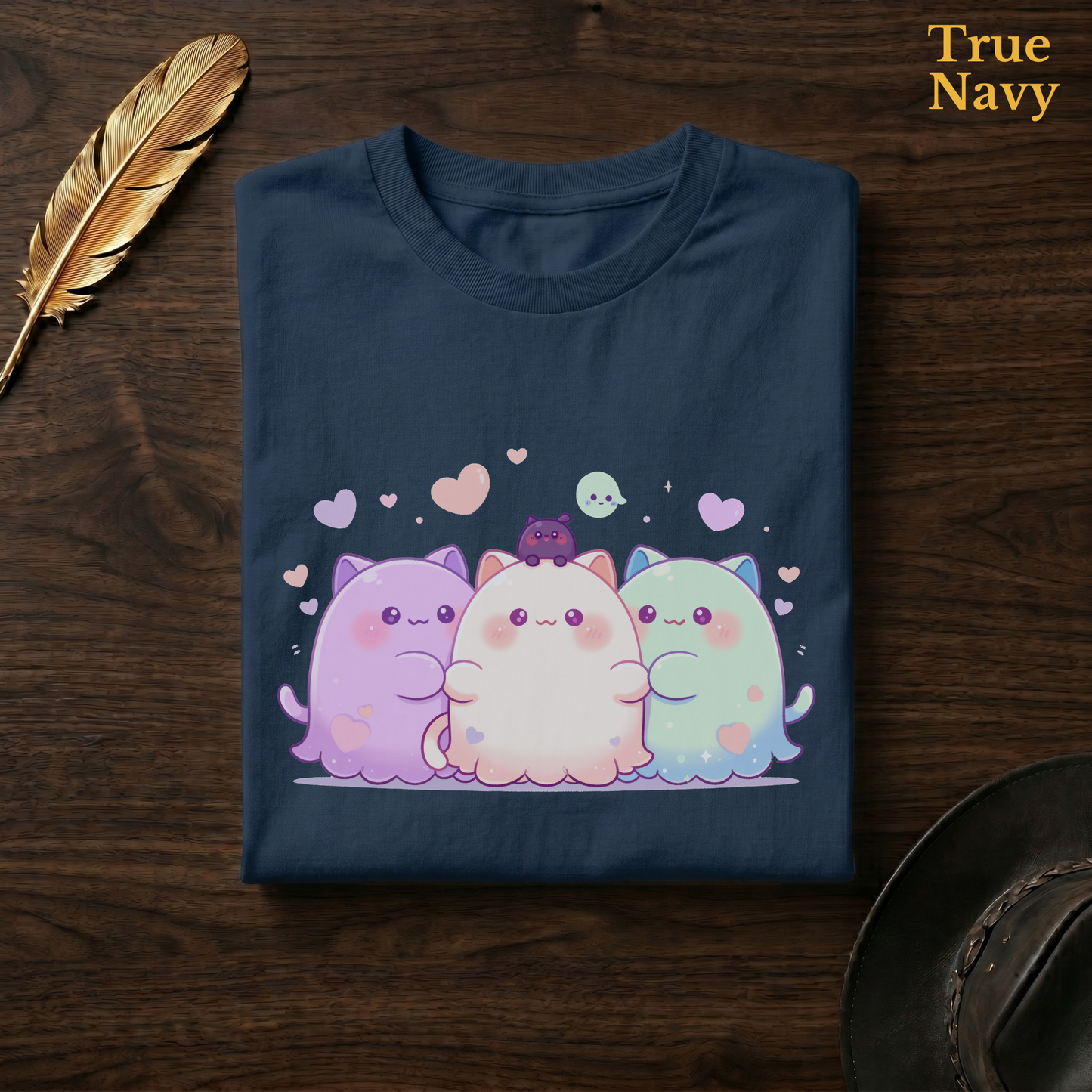 Best friend ghost tee in True Navy, a deep navy garment-dyed shirt folded flat on dark walnut wood beside a gold feather and a leather hat, the pastel cat-ghost trio design on the chest, labelled True Navy.
