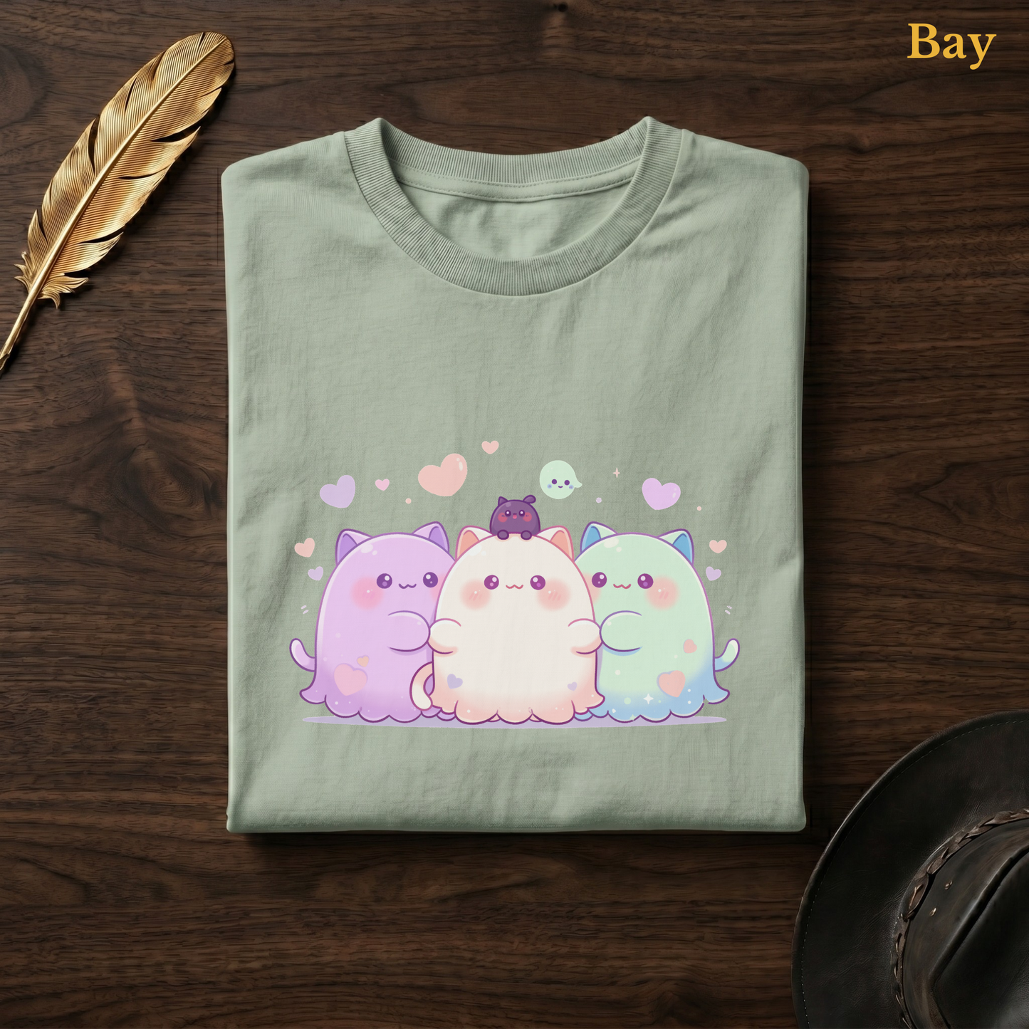 Ghoul Friends tee in Bay, a soft sage-green garment-dyed shirt folded flat on dark walnut wood beside a gold feather and a leather hat, the chubby cat-ghost trio design printed on the chest, labelled Bay.
