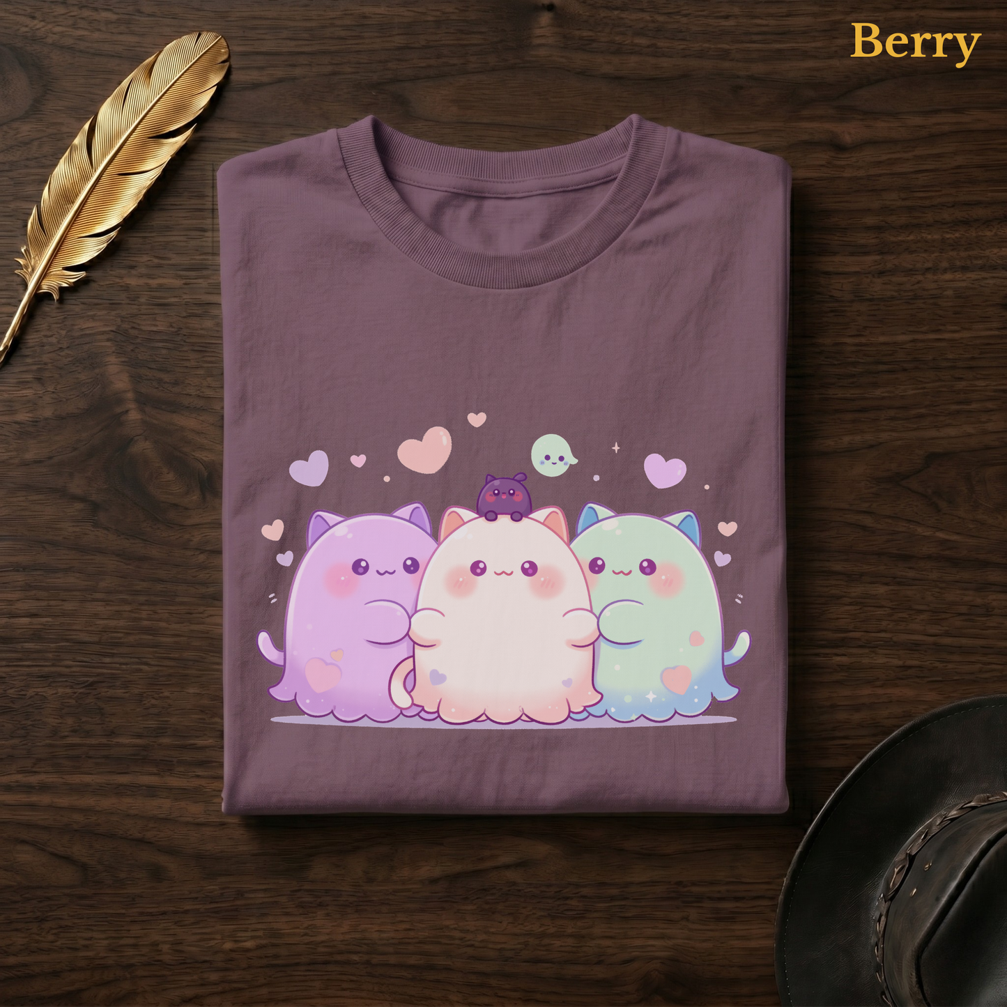 Ghoul Friends tee in Berry, a muted mauve-plum garment-dyed shirt folded flat on dark walnut wood beside a gold feather and a leather hat, the kawaii blob ghost design on the chest, labelled Berry.
