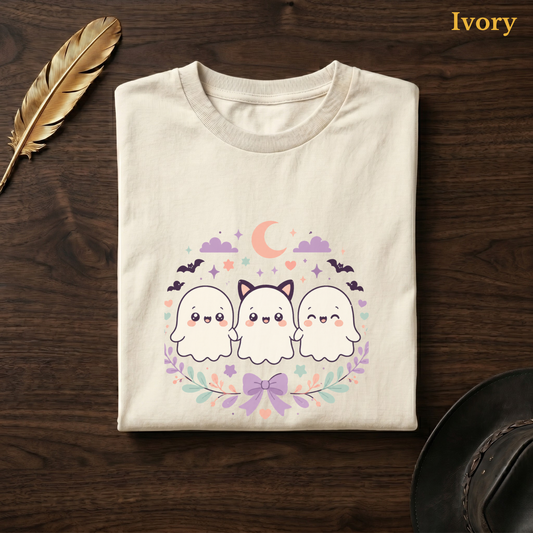 Cute ghost shirt in Ivory, folded flat on dark walnut wood beside a gold feather and a leather hat, the kawaii ghost trio, bats, and floral wreath design on the chest, labelled Ivory.