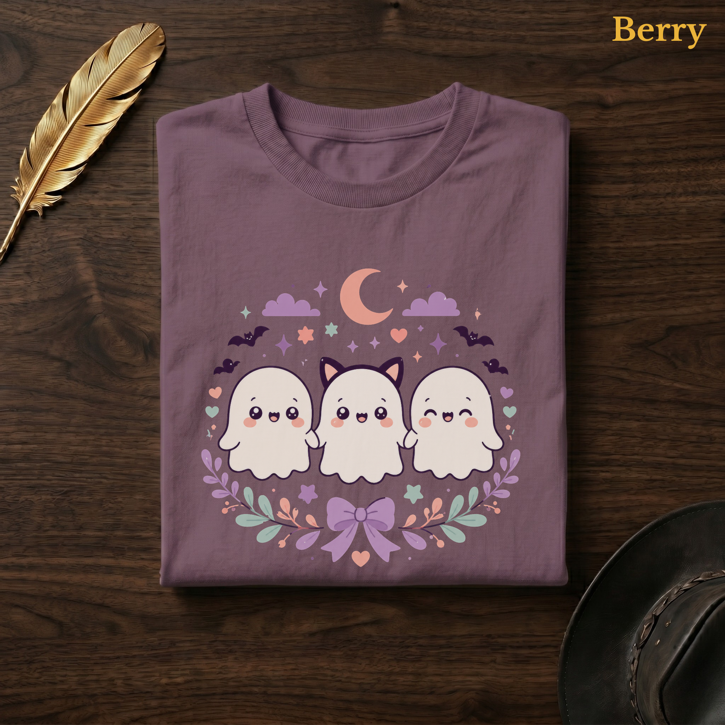 Ghoul Friends tee in Berry, a muted mauve-plum garment-dyed shirt folded flat on dark walnut wood beside a gold feather and a leather hat, the cute ghost squad design on the chest, labelled Berry.