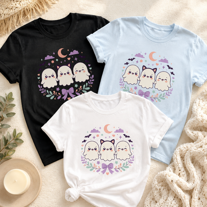 Kawaii ghost tee shown in Black, Chambray, and White laid flat on a cream rug with a woven mat, a candle, and a knit throw, the White tee knotted at the hem, the Ghoul Friends design on each chest.