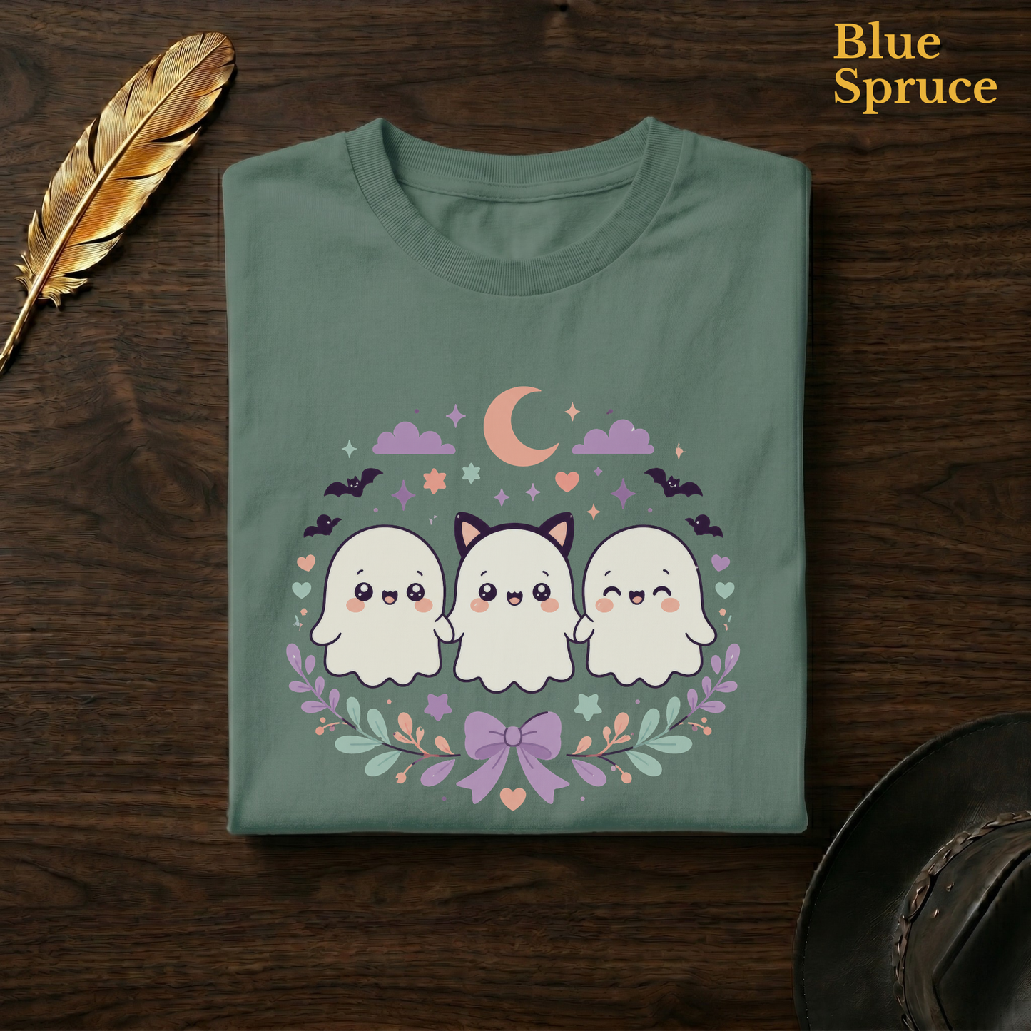 Pastel ghost shirt in Blue Spruce, a deep teal-green garment-dyed shirt folded flat on dark walnut wood beside a gold feather and a leather hat, the kawaii ghost squad design on the chest, labelled Blue Spruce.