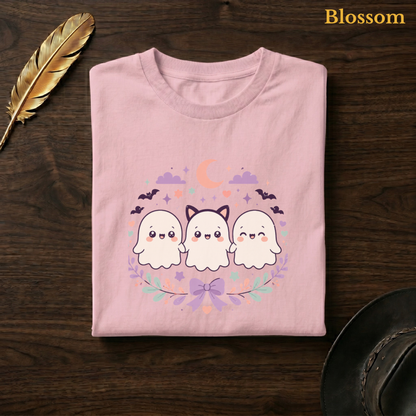 Ghoul Friends tee in Blossom, a soft pastel pink garment-dyed shirt folded flat on dark walnut wood beside a gold feather and a leather hat, the kawaii ghost design on the chest, labelled Blossom.