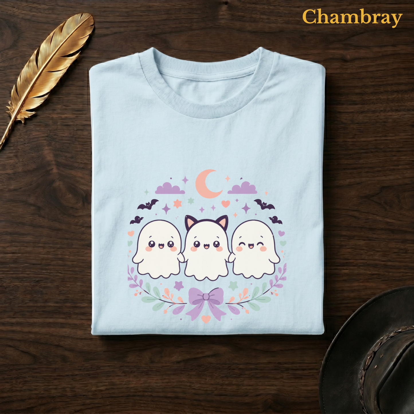 Ghoul Friends tee in Chambray, a soft pale blue garment-dyed shirt folded flat on dark walnut wood beside a gold feather and a leather hat, the cat-ear ghost trio design on the chest, labelled Chambray.