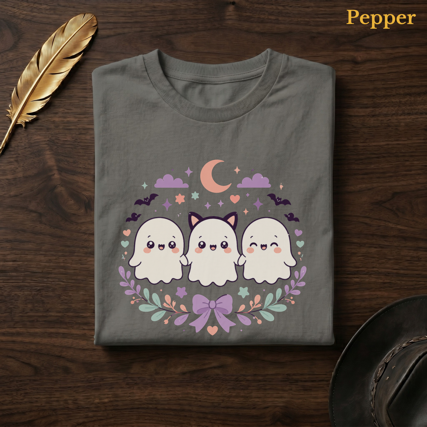 Spooky cute tshirt in Pepper, a warm grey-charcoal garment-dyed shirt folded flat on dark walnut wood beside a gold feather and a leather hat, the Ghoul Friends design centred on the chest, labelled Pepper.